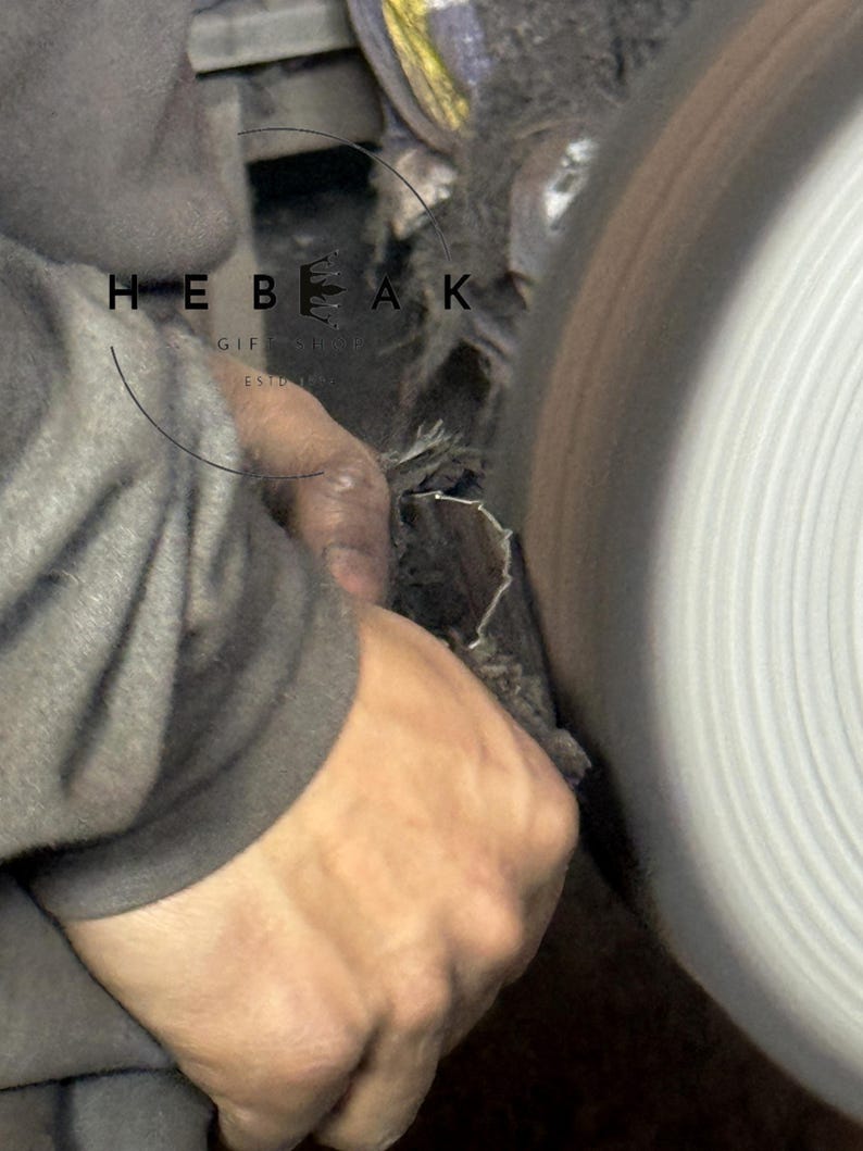 May include: Close-up of a craftsman working on a metal object with a grinding wheel. The image shows the hands of the artisan holding the metal piece against the spinning wheel. The text "HEBEAK GIFT SHOP ESTD" is visible.