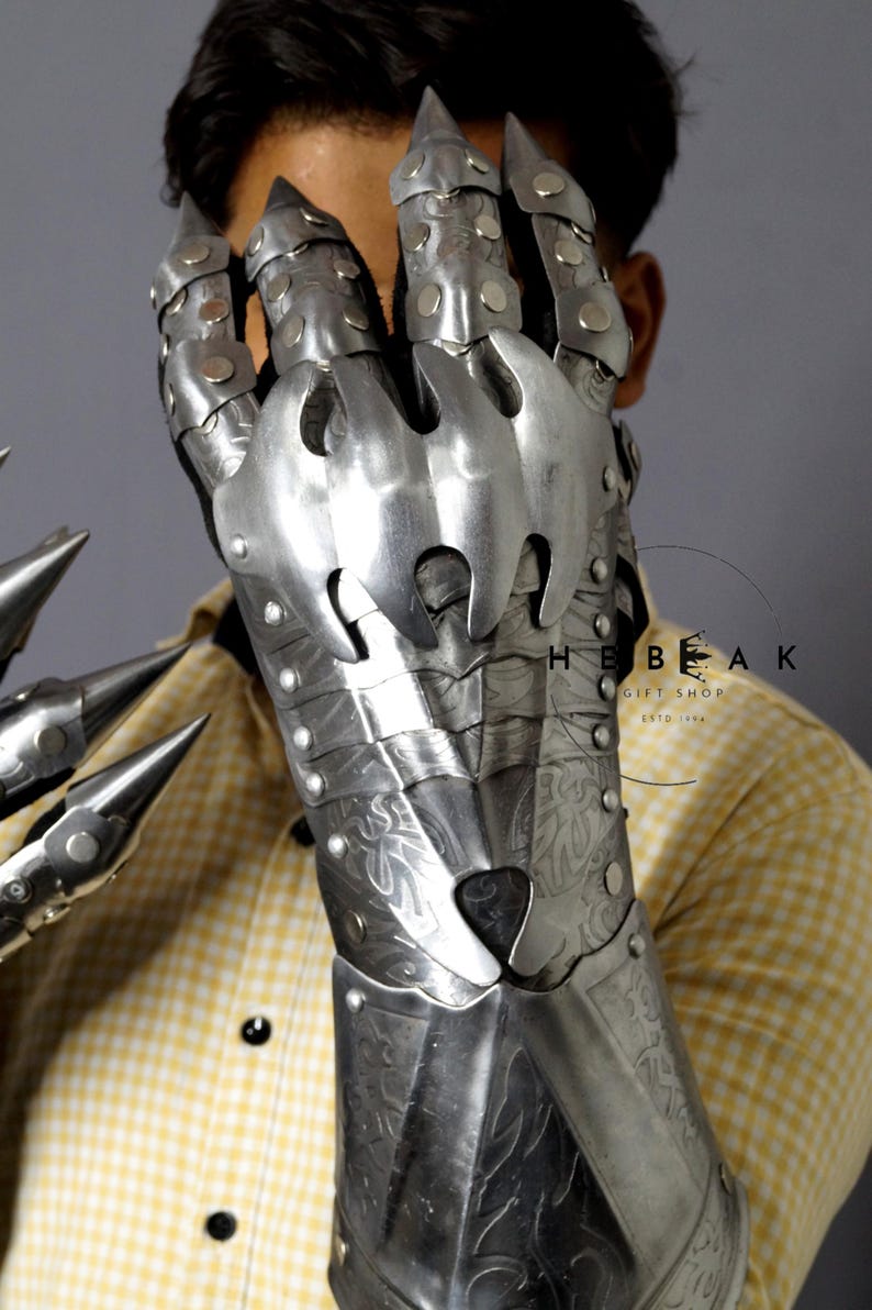 K&ouml;nnte beinhalten: Ein Paar silberne Metallhandschuhe mit krallenartigen Fingern. Die Handschuhe sind mit komplizierten Mustern verziert und haben eine strukturierte Oberfl&auml;che. Die Handschuhe werden von einer Person getragen, aber das Gesicht der Person ist nicht sichtbar.