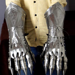 K&ouml;nnte beinhalten: Ein Paar silberne Metallhandschuhe mit Fingerklauen und dekorativen Gravuren. Die Handschuhe werden von einer Person getragen, aber der Fokus liegt auf den Handschuhen selbst.