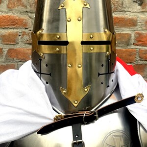May include: A silver knight's helmet with a gold cross design. The helmet has a pointed top and a visor with eye holes. The helmet is worn over a suit of armor with a leather strap.