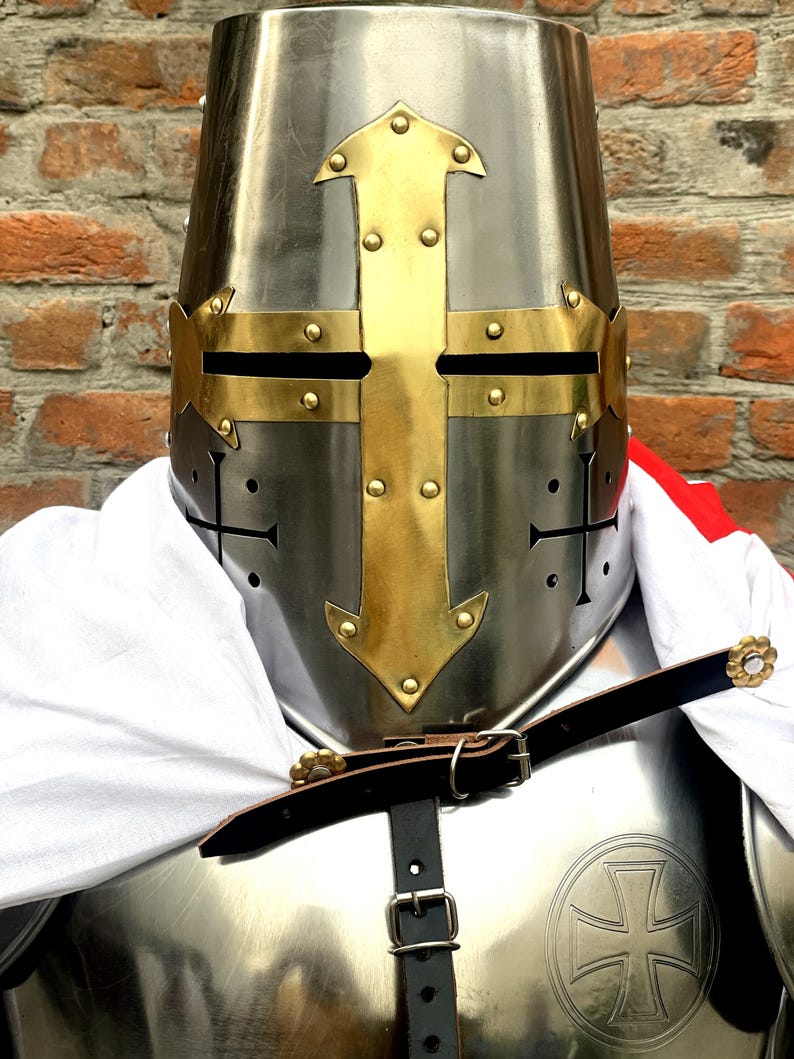 May include: A silver knight's helmet with a gold cross design. The helmet has a pointed top and a visor with eye holes. The helmet is worn over a suit of armor with a leather strap.
