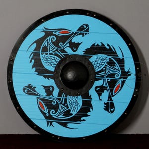 Könnte beinhalten: Ein runder Holzschild mit einem schwarzen Metallrand und einem blauen Hintergrund. Der Schild ist mit einem schwarzen und weißen Design aus zwei ineinander verschlungenen Drachen mit roten Augen verziert.
