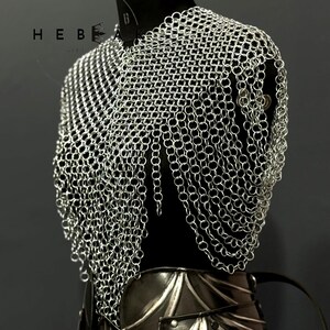 May include: A silver chainmail shirt displayed on a black mannequin. The chainmail is made of small, interlocking metal rings. The mannequin also displays a black leather and metal armor piece. The brand name "HEBE" is visible.