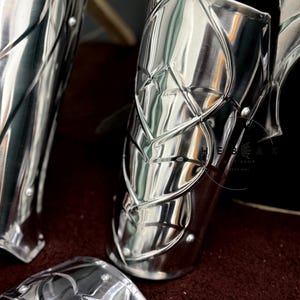 May include: Close-up of three polished silver metal arm guards with intricate designs. The guards are made of metal plates with a raised pattern. The guards are arranged on a dark red surface.