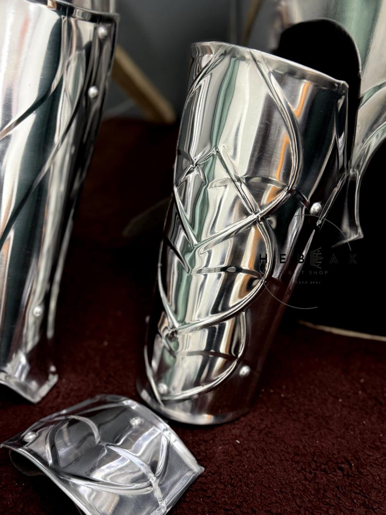 May include: Close-up of three polished silver metal arm guards with intricate designs. The guards are made of metal plates with a raised pattern. The guards are arranged on a dark red surface.