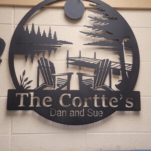 May include: Black metal wall art featuring a lakeside scene with two Adirondack chairs, a dock, and trees. The text "The Cortte's Dan and Sue" is displayed below the scene, making it a personalized home decor item.