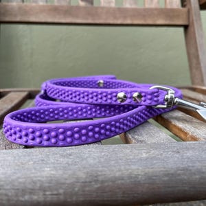 May include: A purple dog leash with a textured, dotted pattern. The leash has a silver metal clasp and rivets. The leash is lying on a wooden chair.