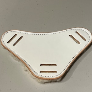 May include: A white leather piece with a curved shape and three pairs of parallel slits. The edges are stitched with a light-colored thread, and the underside has a tan, fuzzy texture. The item appears to be a protective leather component.