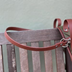 Op de afbeelding: Een bruine leren riem buigt over een houten bank, verbonden met een groen-bruine tas. De tas heeft zilveren hardware en bruine lederen details. De achtergrond is een lichtgroene muur.