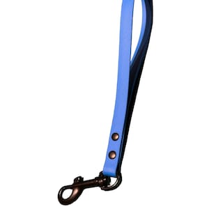 Puede incluir: Una correa de muñeca de cuero azul con un cierre de metal negro y remaches. La correa está diseñada para rodear la muñeca y sujetarse a una bolsa u otro artículo. La correa mide aproximadamente 25 cm de largo.