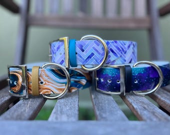 May include: Three dog collars with unique designs. One features a blue and purple chevron pattern, another has a marbled effect in teal and gold, and the third showcases a galaxy-like design in blue and purple. Each collar has a metal buckle and D-ring.