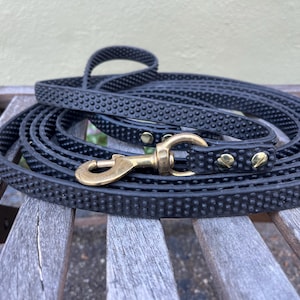 May include: A black dog leash with a textured, dotted pattern is coiled on a wooden surface. The leash features a brass-colored clasp and rivets. The handle is looped, and the leash appears durable and ready for use.