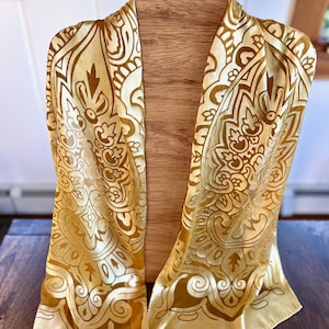 May include: A gold-colored scarf with an intricate, repeating pattern in a darker gold hue. The scarf is draped over a wooden form, showcasing its flowing design and detailed embellishments. The scarf's edges are finished with a subtle border.
