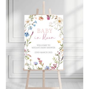 May include: A white sign with a floral wreath border. The sign says "Baby in Bloom" in pink and black lettering. The text below says "Welcome to Megan's Baby Shower 22nd March 2025".