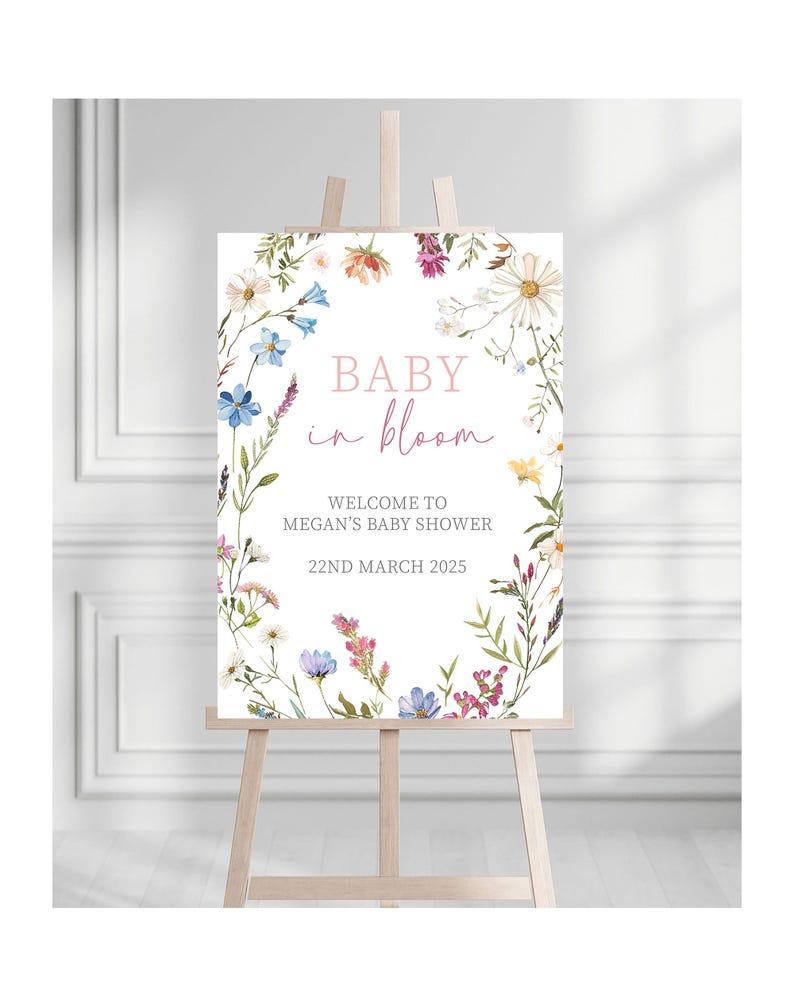 May include: A white sign with a floral wreath border. The sign says "Baby in Bloom" in pink and black lettering. The text below says "Welcome to Megan's Baby Shower 22nd March 2025".