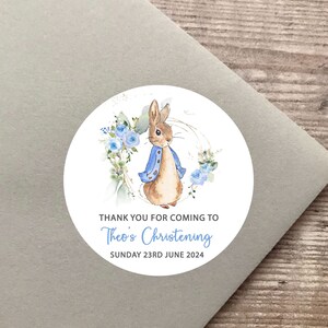 Op de afbeelding: Ronde witte sticker met een aquarel-illustratie van Peter Rabbit, die een blauw jasje draagt en omringd is door blauwe bloemen. De tekst "Bedankt voor uw komst naar de doop van Theo Zondag 23 juni 2024" staat onder de illustratie.