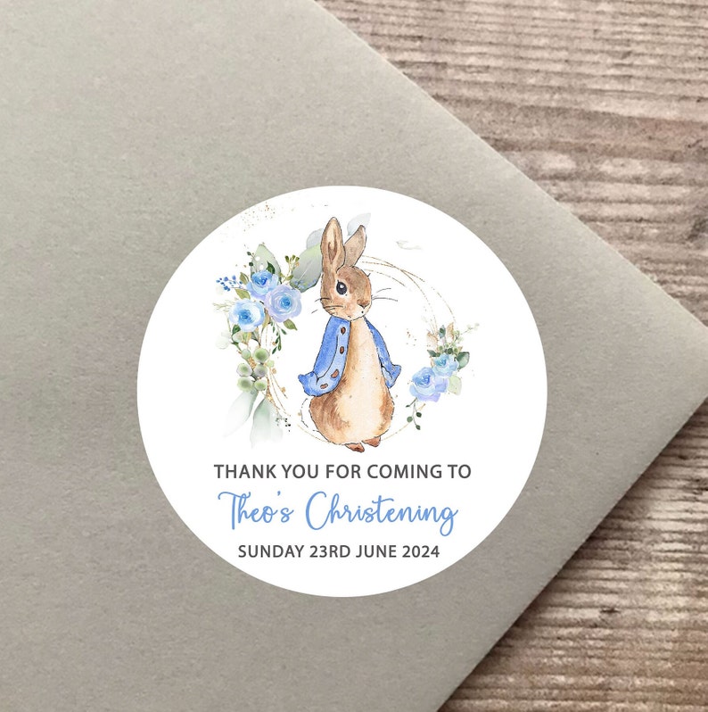 Op de afbeelding: Ronde witte sticker met een aquarel-illustratie van Peter Rabbit, die een blauw jasje draagt en omringd is door blauwe bloemen. De tekst "Bedankt voor uw komst naar de doop van Theo Zondag 23 juni 2024" staat onder de illustratie.