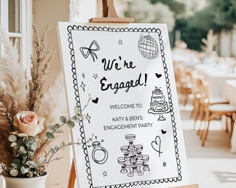 May include: A white sign on a wooden easel with the text "We're Engaged!" in black script. The sign includes illustrations of a bow, disco ball, cake, ring, champagne glasses, and hearts. The sign welcomes guests to Katy & Ben's engagement party.