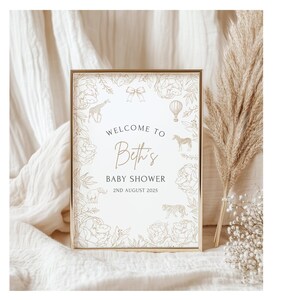 May include: A framed baby shower sign with a gold border. The sign reads "Welcome to Beth's Baby Shower 2nd August 2025". The sign is decorated with floral and animal illustrations in a gold color.