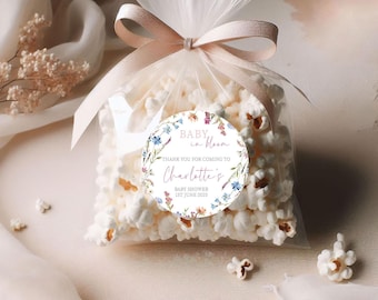 May include: A clear cellophane bag filled with popcorn, tied with a light pink ribbon. A round sticker with floral accents and the text "Baby in Bloom" and "Thank you for coming to Charlotte's Baby Shower 1st June 2025" is attached to the bag.