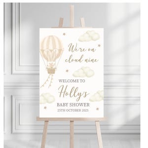 May include: A baby shower welcome sign on an easel. The sign features a hot air balloon with a teddy bear, clouds, and stars. Text reads "We're on cloud nine", "Welcome to Holly's", and "Baby Shower 25th October 2025".