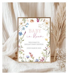 May include: A white sign with a floral border and the text "Baby in bloom" in pink lettering. The sign also includes the text "Welcome to Megan's Baby Shower" and "22nd March 2025".