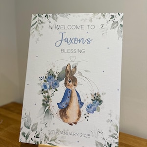 May include: A white welcome sign with a watercolor design of blue flowers and green leaves. The sign features a cartoon rabbit wearing a blue jacket and the text "Welcome to Jaxon's Blessing 19th January 2025".