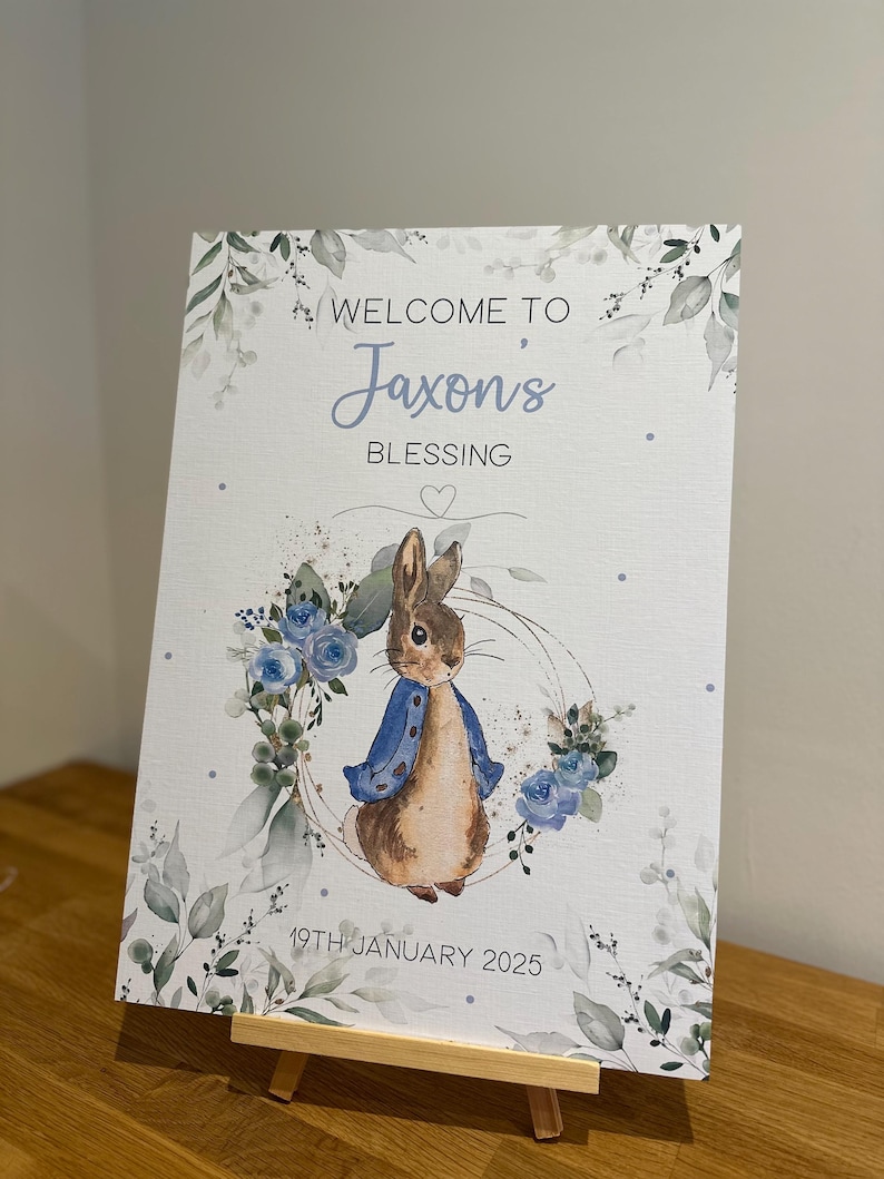May include: A white welcome sign with a watercolor design of blue flowers and green leaves. The sign features a cartoon rabbit wearing a blue jacket and the text "Welcome to Jaxon's Blessing 19th January 2025".