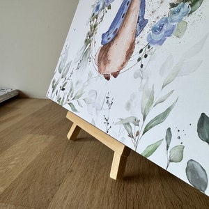 May include: A watercolor painting of a rabbit wearing a blue jacket, surrounded by blue flowers and green leaves. The painting is on a small wooden easel.