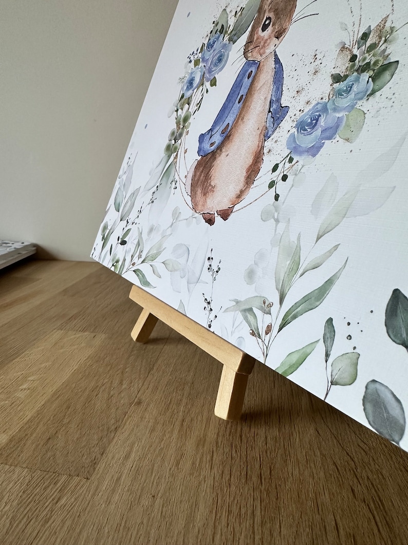 May include: A watercolor painting of a rabbit wearing a blue jacket, surrounded by blue flowers and green leaves. The painting is on a small wooden easel.