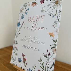 May include: A white card with a floral border and the text "BABY in bloom" in pink script. The card also includes the text "WELCOME TO HOLLY'S BABY SHOWER 27TH APRIL 2025".