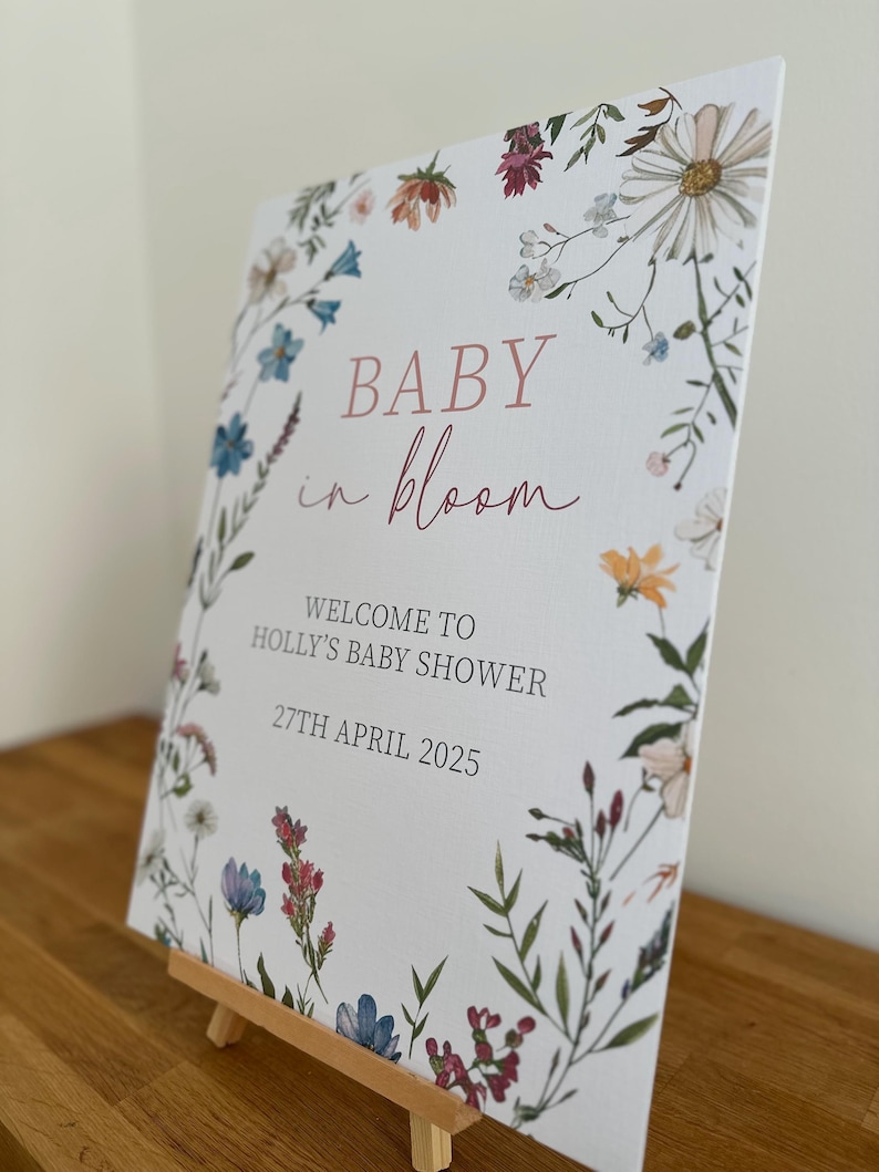 May include: A white card with a floral border and the text "BABY in bloom" in pink script. The card also includes the text "WELCOME TO HOLLY'S BABY SHOWER 27TH APRIL 2025".