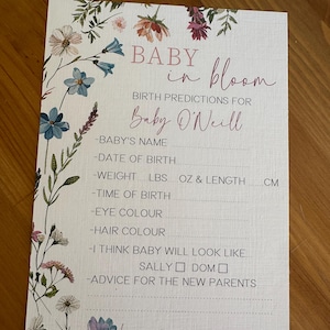 May include: A baby shower game card with the text "BABY in bloom" and "Birth Predictions for Baby O'Neill". The card is decorated with floral illustrations in shades of blue, pink, and white. It includes prompts for baby's name, date of birth, weight, and more.