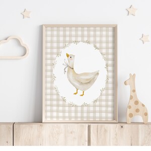 May include: A framed print with a beige and white gingham pattern. The print features a watercolor illustration of a white goose with a yellow beak and feet, surrounded by a wreath of green leaves. The goose is standing on a white background.