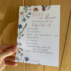 May include: A baby shower prediction card with floral accents. The card reads "Baby in Bloom" and includes prompts for baby's name, birth details, and advice for parents. The card is held against a wooden background.