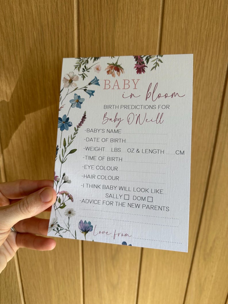 May include: A baby shower prediction card with floral accents. The card reads "Baby in Bloom" and includes prompts for baby's name, birth details, and advice for parents. The card is held against a wooden background.