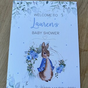 May include: A baby shower invitation with a blue and white watercolor design featuring a rabbit with a blue jacket and a wreath of blue flowers. The text reads "Welcome to Lauren's Baby Shower" and "23rd March 2024".