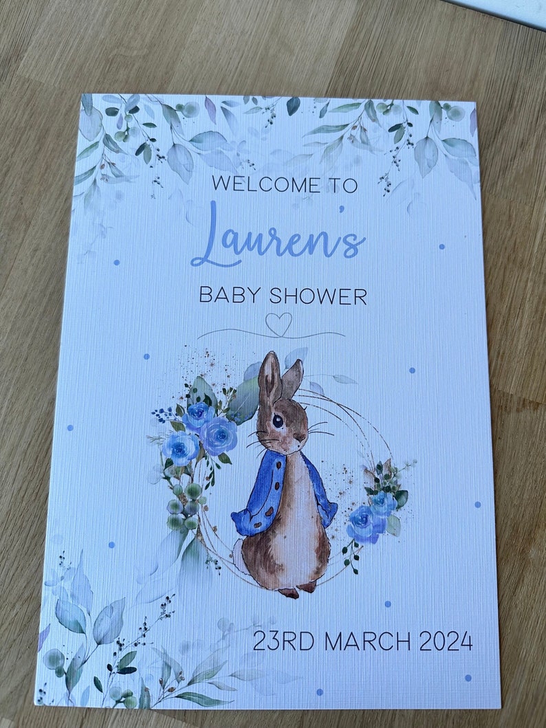 May include: A baby shower invitation with a blue and white watercolor design featuring a rabbit with a blue jacket and a wreath of blue flowers. The text reads "Welcome to Lauren's Baby Shower" and "23rd March 2024".