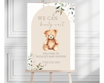 May include: A beige welcome sign with a brown teddy bear and green foliage. The sign reads "We can barely wait" and "Welcome to Natalie's Baby Shower 27th July 2024".