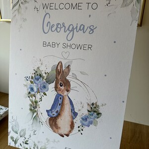 May include: A white sign with a watercolor design of leaves and flowers. The sign says "Welcome to Georgia's Baby Shower" with a heart and the date "5th May 2024". A cartoon rabbit wearing a blue jacket is in the center of the sign.