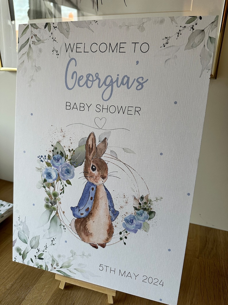 May include: A white sign with a watercolor design of leaves and flowers. The sign says "Welcome to Georgia's Baby Shower" with a heart and the date "5th May 2024". A cartoon rabbit wearing a blue jacket is in the center of the sign.