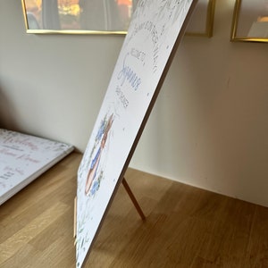 May include: A white baby shower sign with a blue bunny illustration and the text "Welcome to,  Sayan's Baby Shower". The sign is propped up on a wooden easel.