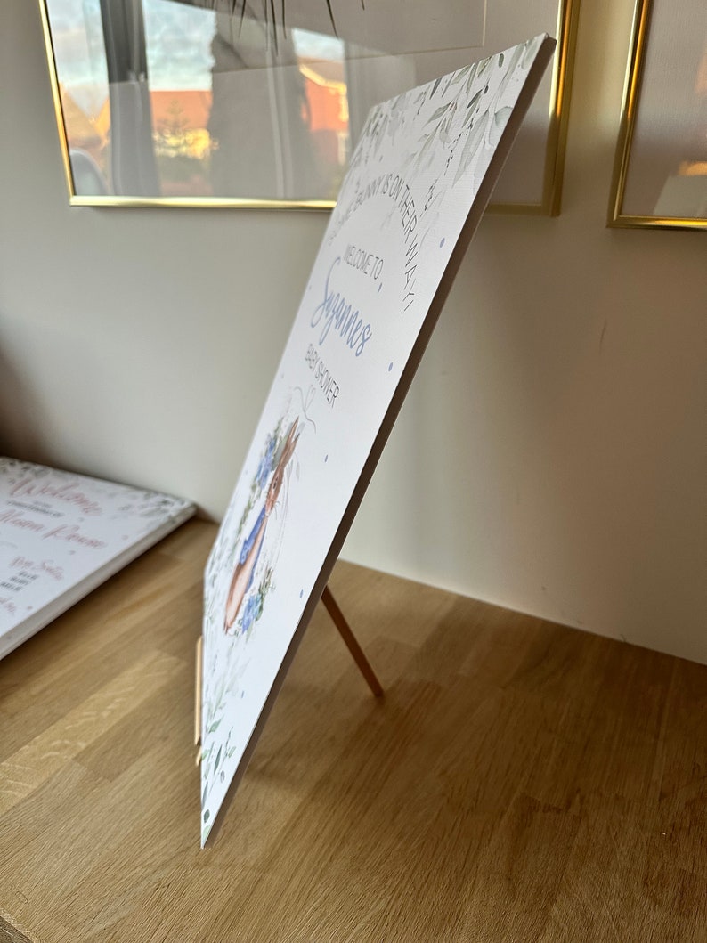 May include: A white baby shower sign with a blue bunny illustration and the text "Welcome to,  Sayan's Baby Shower". The sign is propped up on a wooden easel.