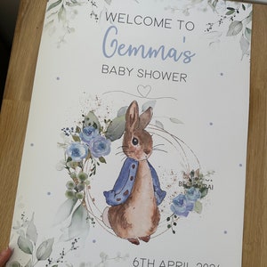 Op de afbeelding: Een wit babyborrelbord met een aquarel-illustratie van een konijn dat een blauw jasje draagt. Het bord zegt "Welkom op Gemma's babyborrel" en "6 april 2024".