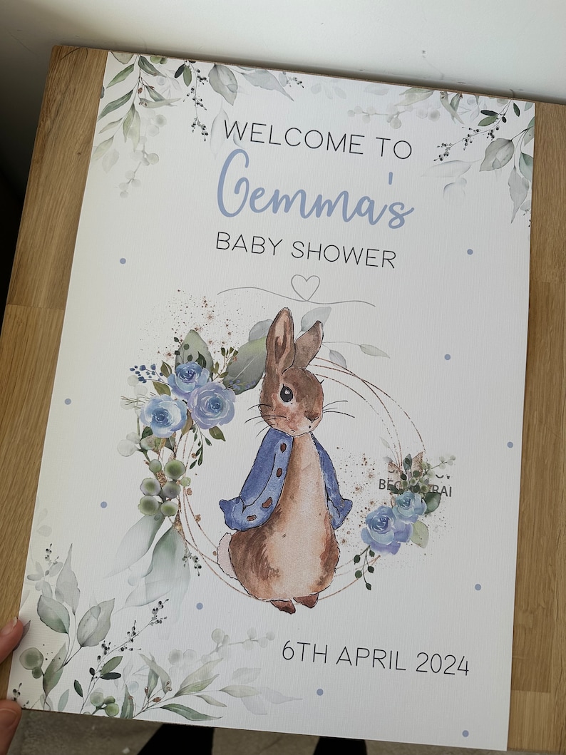 Op de afbeelding: Een wit babyborrelbord met een aquarel-illustratie van een konijn dat een blauw jasje draagt. Het bord zegt "Welkom op Gemma's babyborrel" en "6 april 2024".