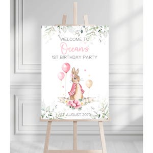 May include: A wooden easel supports a sign welcoming guests to "Ocean's 1st Birthday Party." The sign features a watercolour illustration of a bunny in a pink jacket, pink balloons, and floral accents. The date "1st August 2025" is also displayed.