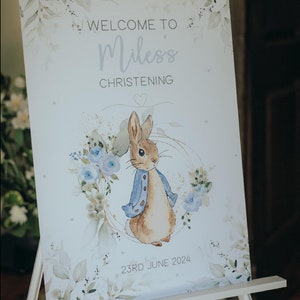 Op de afbeelding: Een wit bord met een aquarelontwerp van blauwe bloemen en groene bladeren. Het bord zegt "Welkom op de doop van Miles" en "23 juni 2024". Een cartoonillustratie van Peter Rabbit in een blauw jasje bevindt zich in het midden van het bord.