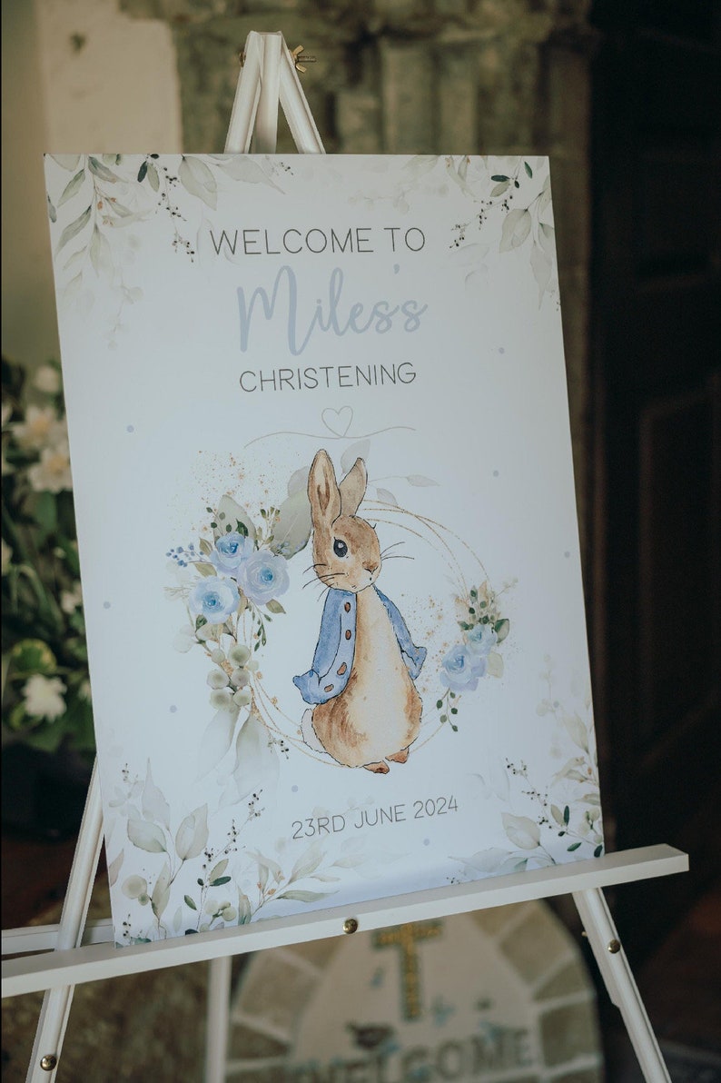 Op de afbeelding: Een wit bord met een aquarelontwerp van blauwe bloemen en groene bladeren. Het bord zegt "Welkom op de doop van Miles" en "23 juni 2024". Een cartoonillustratie van Peter Rabbit in een blauw jasje bevindt zich in het midden van het bord.