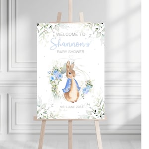 May include: Welcome to Shannon's Baby Shower sign with a watercolor illustration of Peter Rabbit in a blue jacket surrounded by blue flowers and greenery. The date 10th June 2023 is printed below the illustration.
