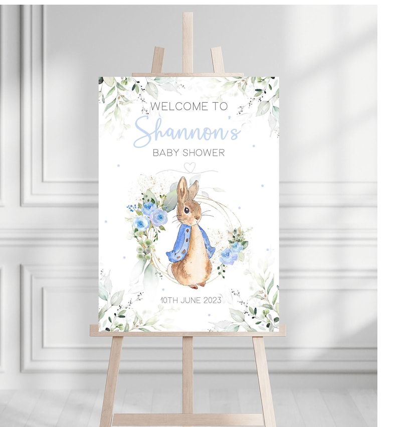 May include: Welcome to Shannon's Baby Shower sign with a watercolor illustration of Peter Rabbit in a blue jacket surrounded by blue flowers and greenery. The date 10th June 2023 is printed below the illustration.